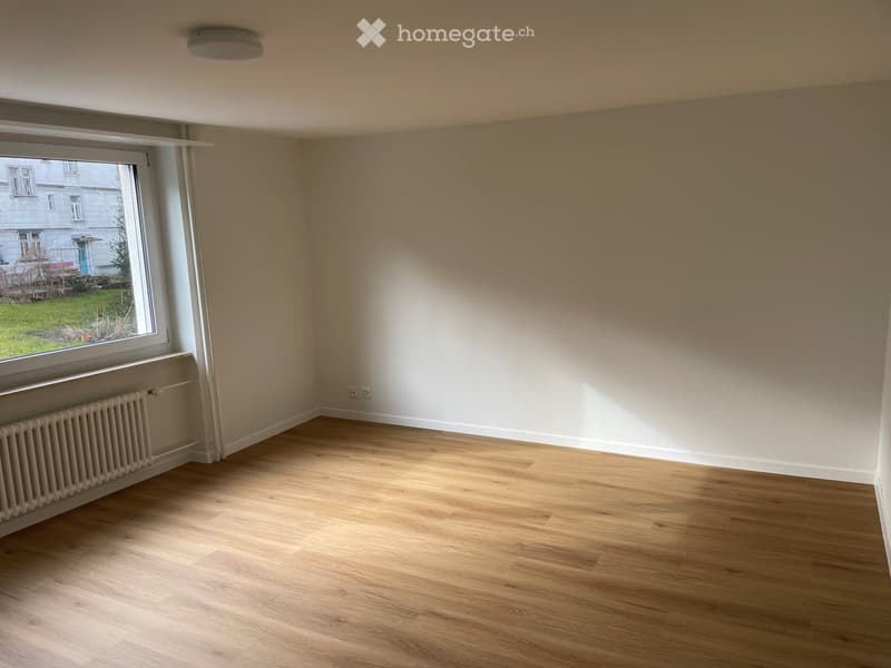 Empty room with wooden flooring, white walls, and a large window with a radiator below it. No furniture or equipment is present.