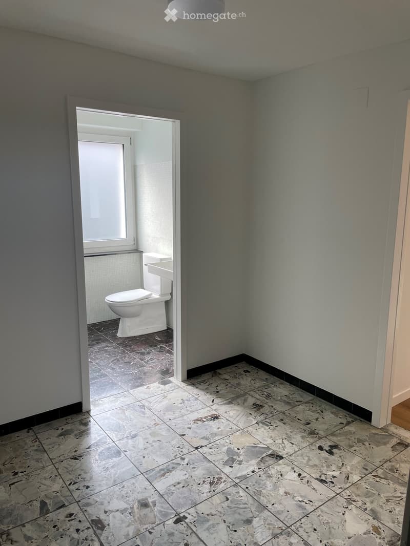 Empty bathroom with marble flooring, white walls, and a toilet visible in an adjacent room through an open doorway.