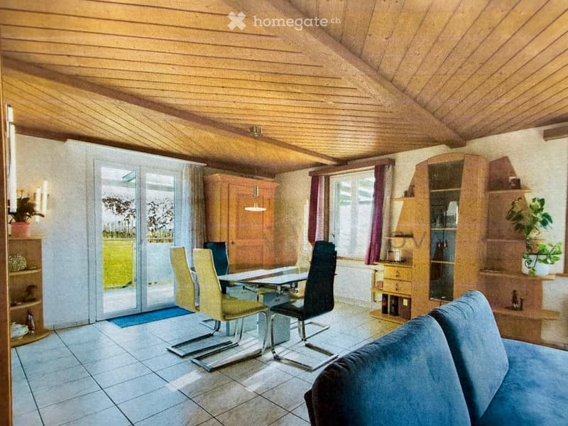 Bright living and dining area with wooden ceiling, tiled floor, large windows, and sliding glass doors leading to a balcony. Features a blue sofa, dining table with chairs, wooden cabinets, and decorative plants.