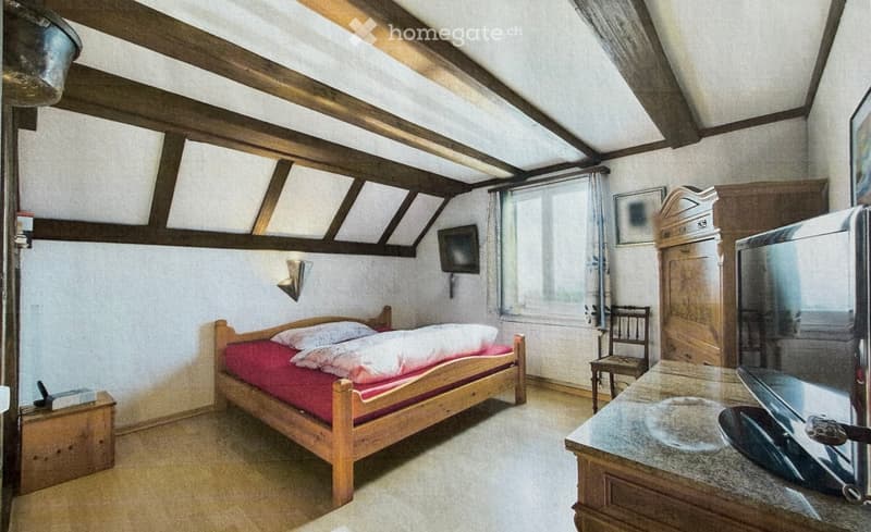 Cozy attic bedroom with wooden beams, a single bed with red bedding, a small wooden chair, a television, and a wooden cabinet.