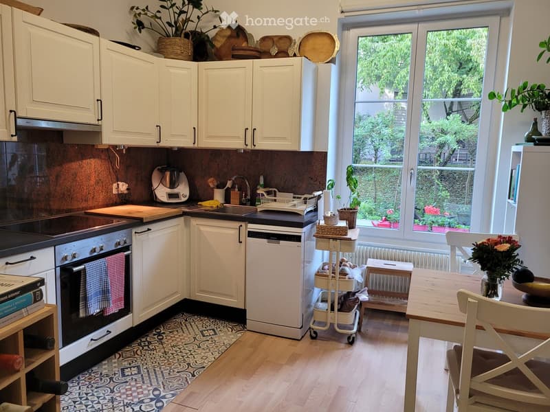 Modern kitchen with white cabinets, stainless steel appliances, and a large window overlooking a garden. Includes an oven, dishwasher, sink, cutting board, blender, cart, dining table, chairs, and decorative items.