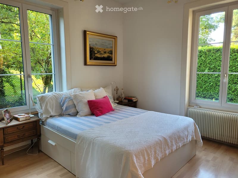 Bright bedroom with large windows, a neatly made bed with pillows and a colorful throw, two nightstands with lamps, and a framed painting on the wall.
