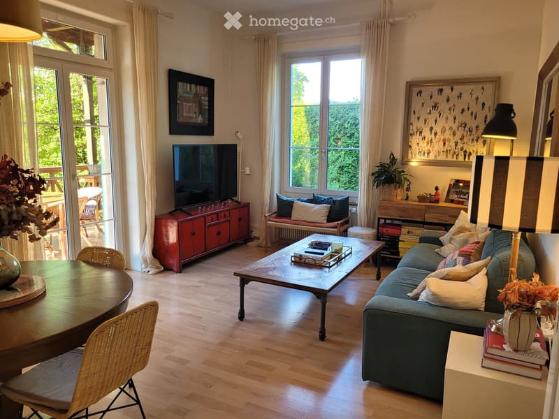 Bright living room with large windows and a view of greenery, featuring a blue sofa, wooden coffee table, dining table with chairs, TV on a red cabinet, and decorative plants.