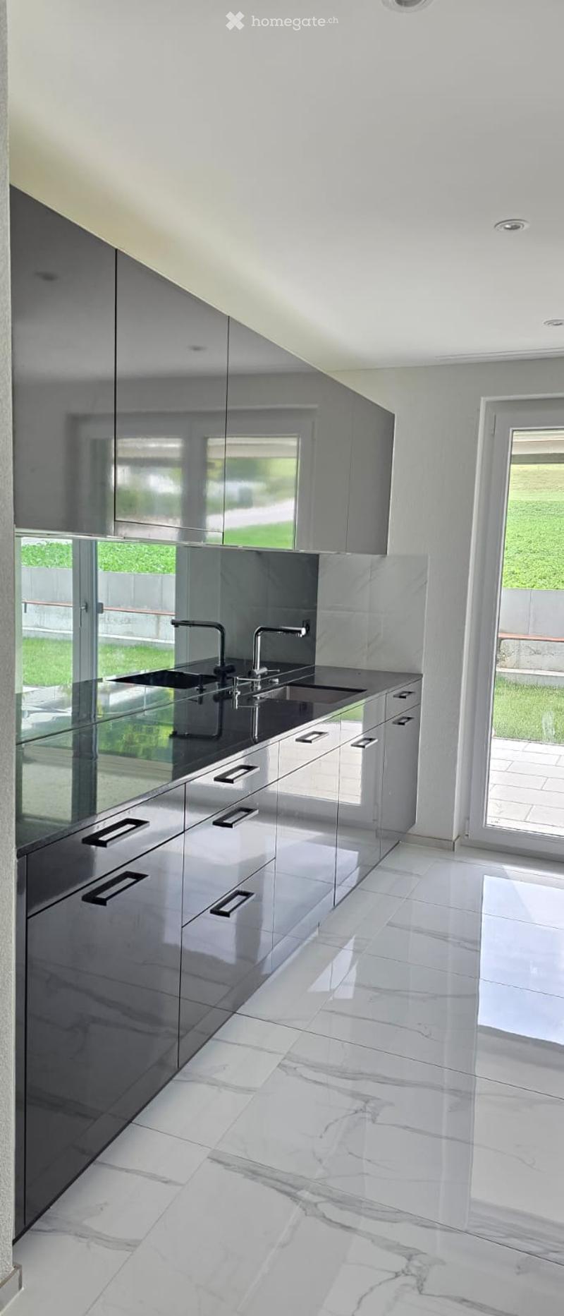 Modern kitchen with sleek, grey cabinets, a double sink, and a marble tiled floor. Large windows provide natural light and a view of the outdoor area.