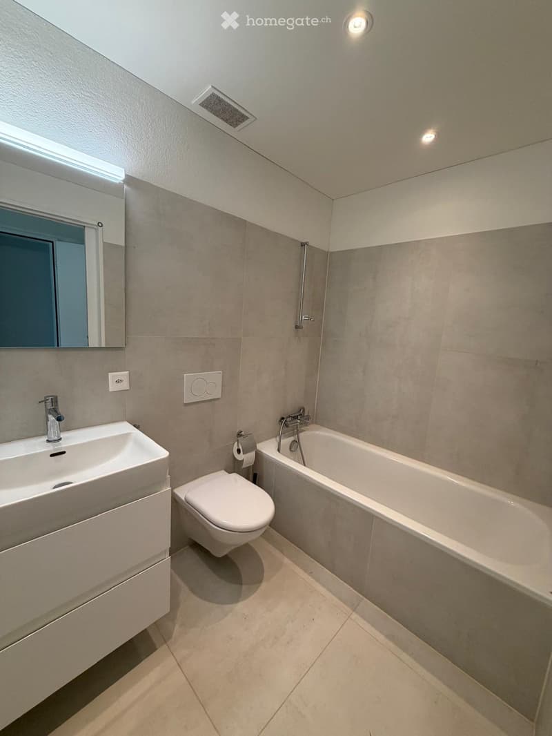 Modern bathroom with a white sink, toilet, bathtub, and mirror. Light grey tiles and a sleek design.