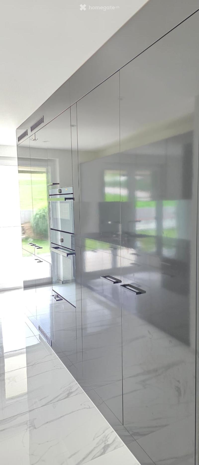 Modern kitchen with sleek, mirrored cabinets and a built-in oven, featuring a polished marble floor and ample natural light from large windows.