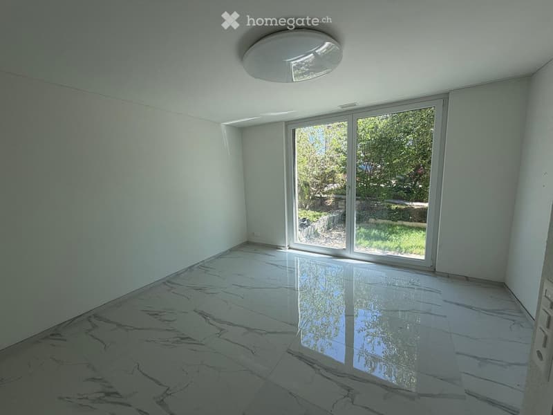 Empty room with white walls, polished marble floor, large sliding glass door leading to a garden, and a modern ceiling light fixture.