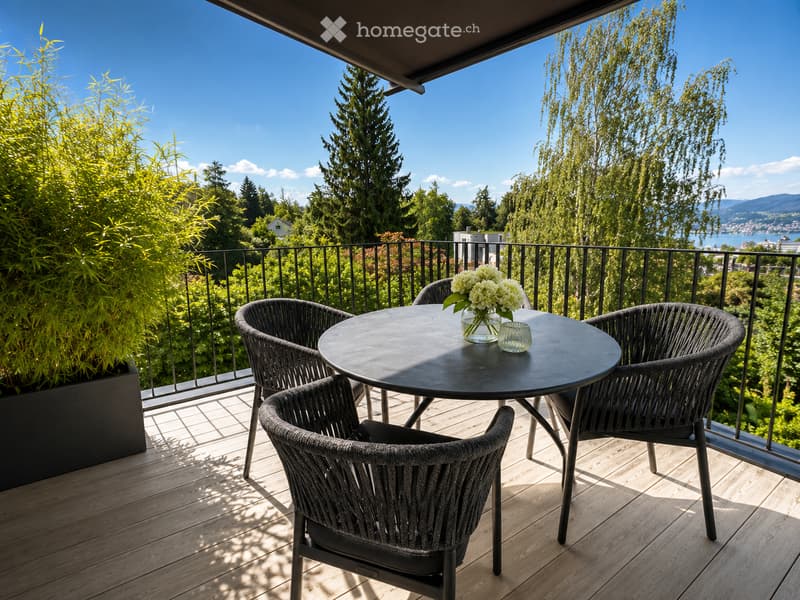 Balcony with wooden decking, round table, four wicker chairs, vase with white flowers, and scenic view of trees and water.