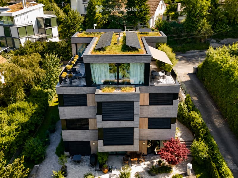 Modern multi-story house with a flat roof, large windows, and solar panels. Features include a balcony with outdoor furniture, a patio area with tables and chairs, and a landscaped garden with potted plants and trees.