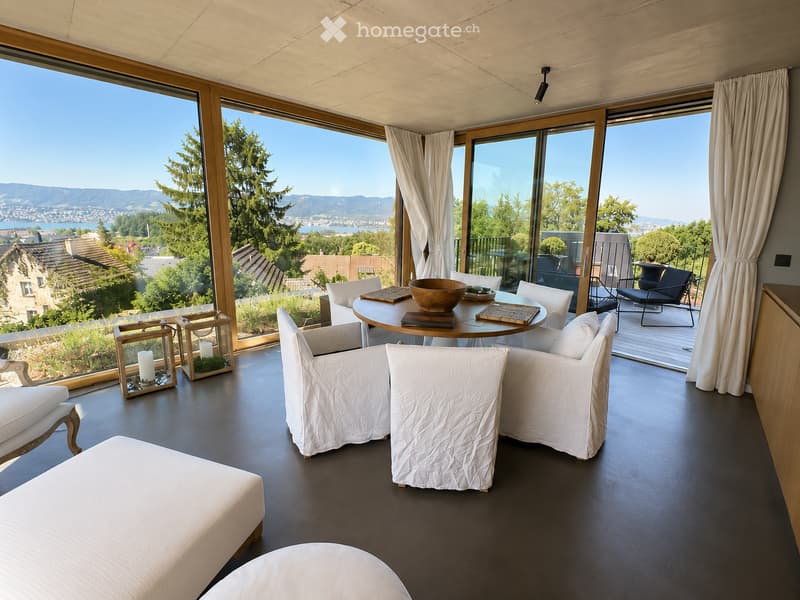 Modern living room with large windows offering a scenic view, featuring white furniture including sofas, chairs, a round table, and decorative candle holders.