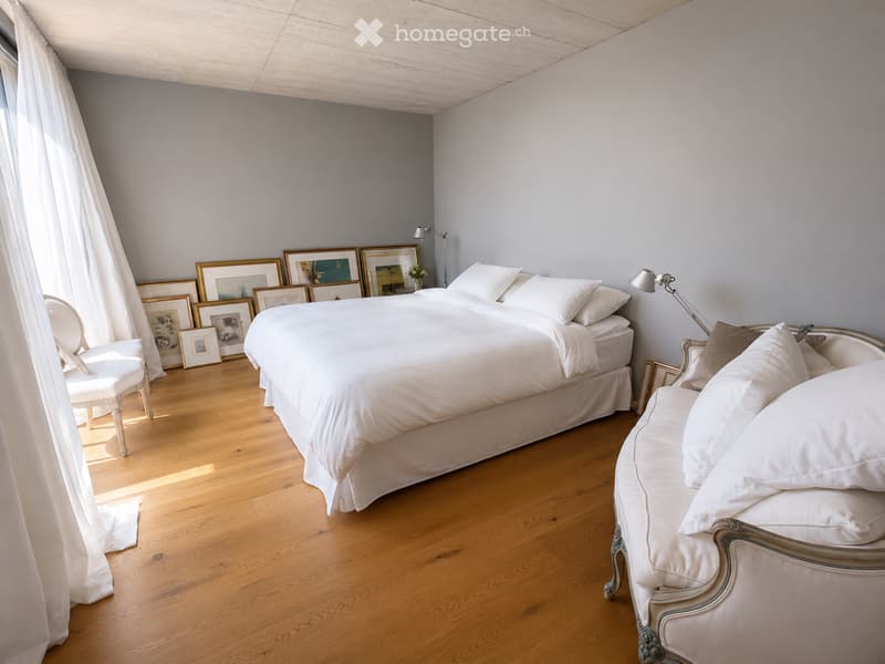 Bright bedroom with wooden flooring, large bed with white bedding, a sofa, a chair, and several picture frames on the wall.