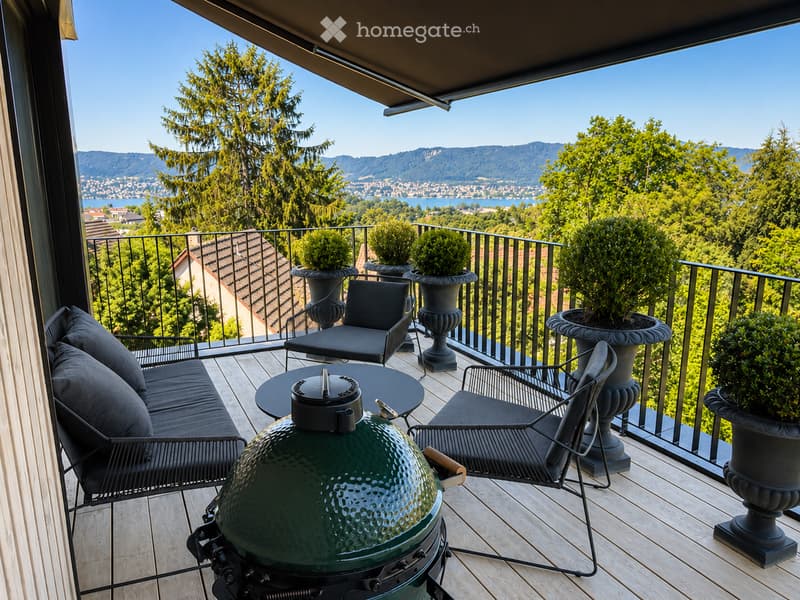 Balcony with wooden decking, black metal railing, and scenic mountain and lake view. Features a green grill, black sofa, chairs, and a small table.