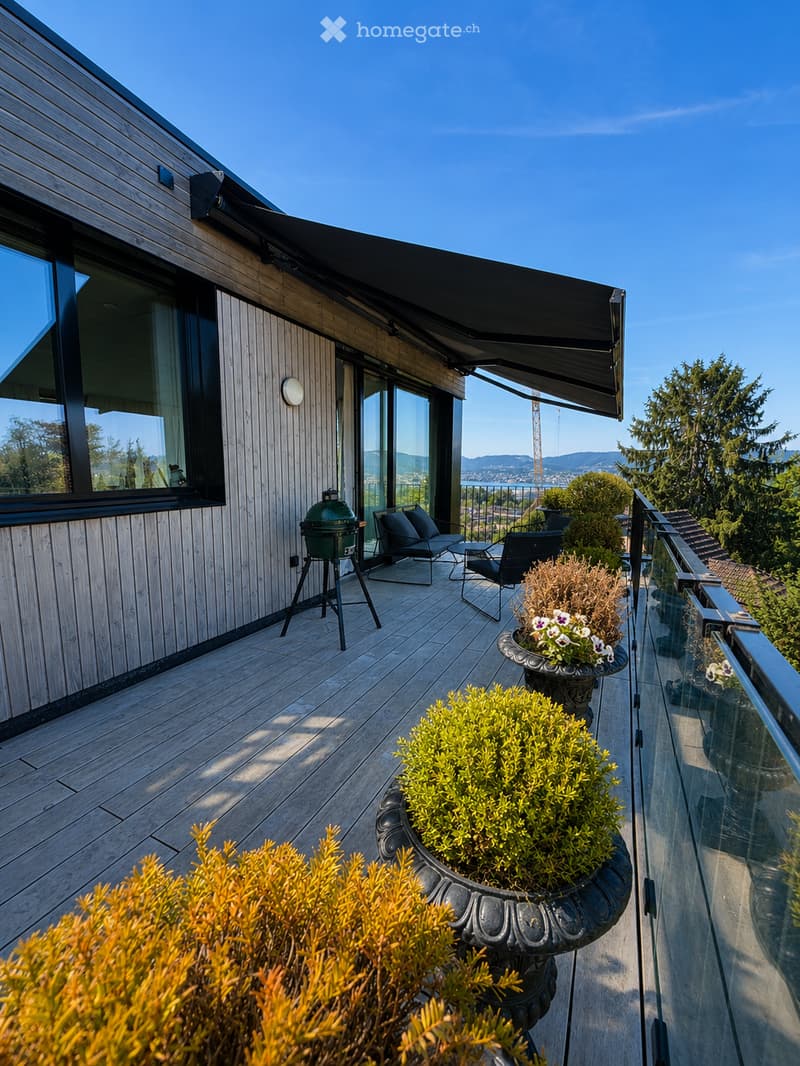 A modern wooden balcony with a glass railing, featuring outdoor seating, a grill, and potted plants, overlooking a scenic view.