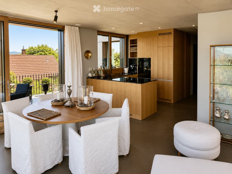 Modern, well-lit room with a round wooden dining table surrounded by white chairs, a white stool, a kitchen island with wooden cabinets, and a glass display cabinet with decorative items. Large windows offer a scenic view of the outdoors.