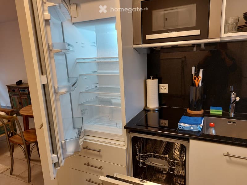 Modern kitchen with stainless steel appliances, including a refrigerator, microwave, dishwasher, and sink. Countertop features a paper towel holder, scissors, and a small table with a chair nearby.