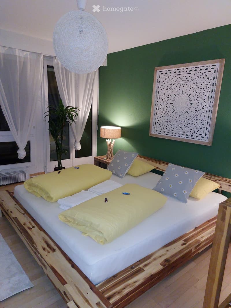 Cozy bedroom with a wooden bed, yellow and blue bedding, nightstand with a table lamp, decorative wall panel, and a large window with white curtains and a plant.