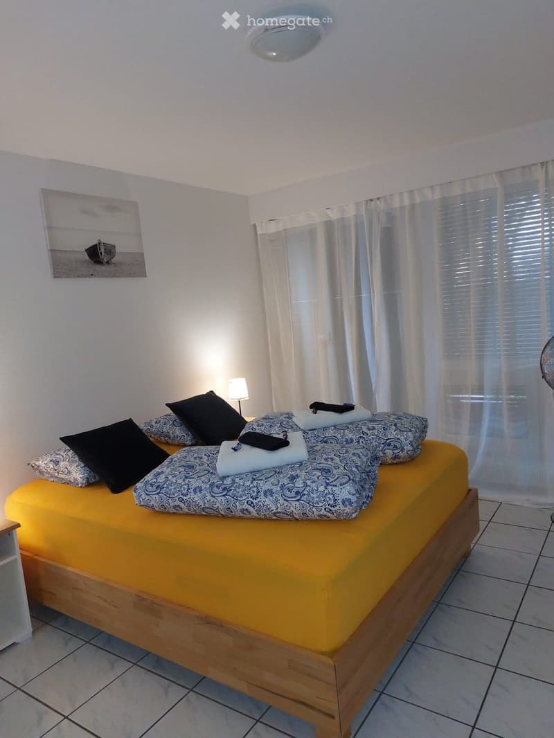 A cozy bedroom with a wooden bed, yellow mattress, patterned bedspread, black and patterned pillows, a bedside lamp, a fan, and a picture on the wall.