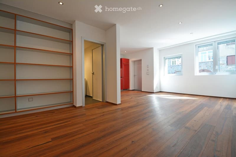 Empty room with wooden flooring, white walls, built-in shelving unit, and large windows letting in natural light.