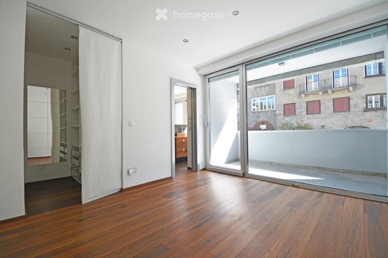 Empty room with wooden flooring, large sliding glass doors leading to a balcony, and a view of a neighboring building.