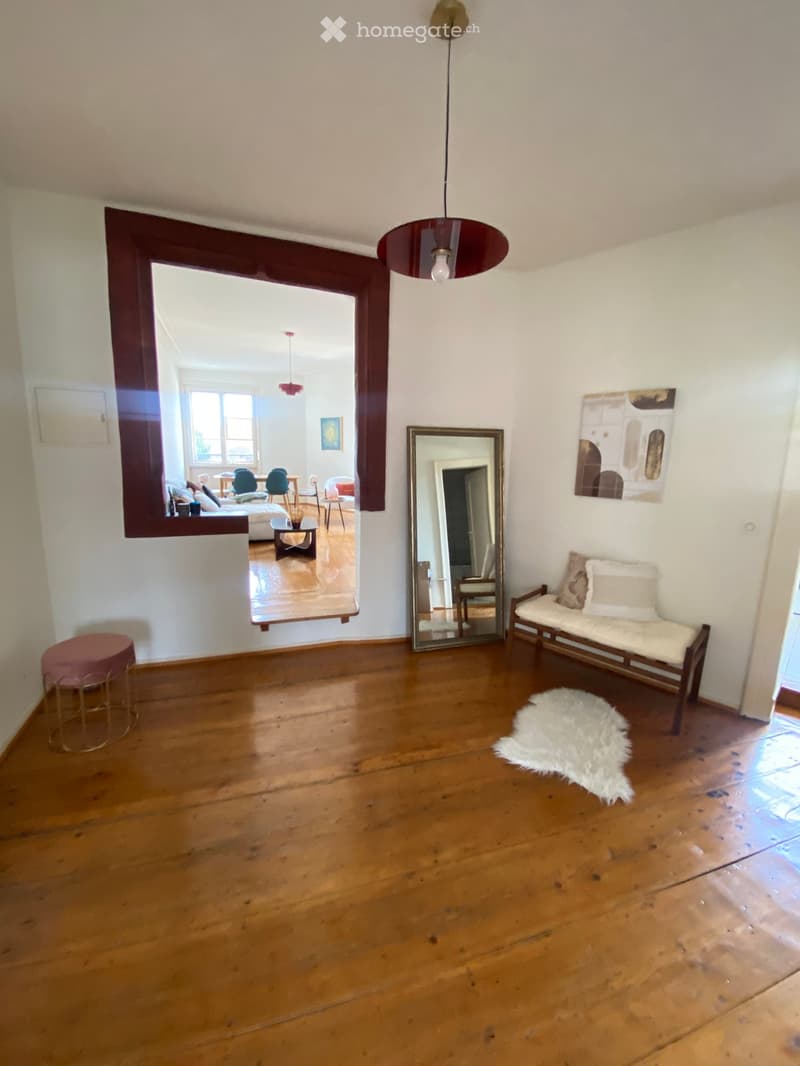 Empty room with wooden flooring, white walls, a large mirror, a hanging lamp, and a small bench with cushions visible in the background.