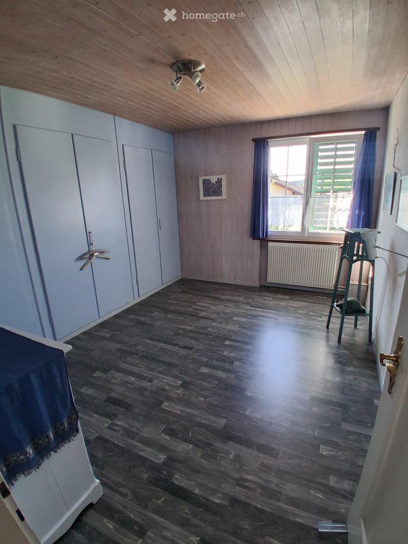 Empty room with wooden flooring, white built-in wardrobes, a window with blue curtains, and a small table with a lamp near the window.