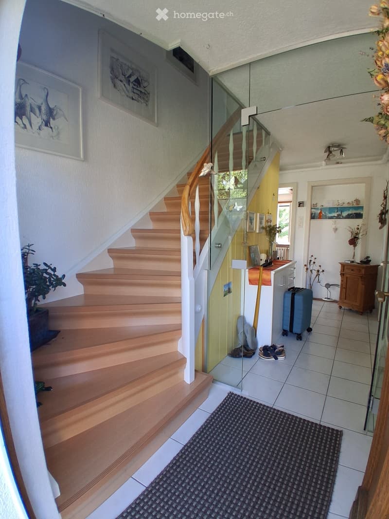 Hallway with wooden staircase, glass railing, and a small entryway area with a suitcase and shoes.