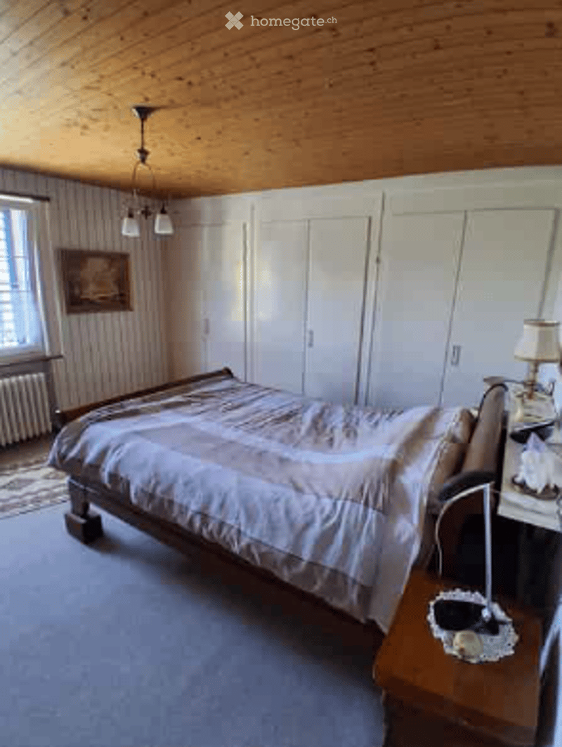 Cozy bedroom with wooden ceiling, large window, bed with patterned bedding, nightstand with lamp, and built-in white cabinets.