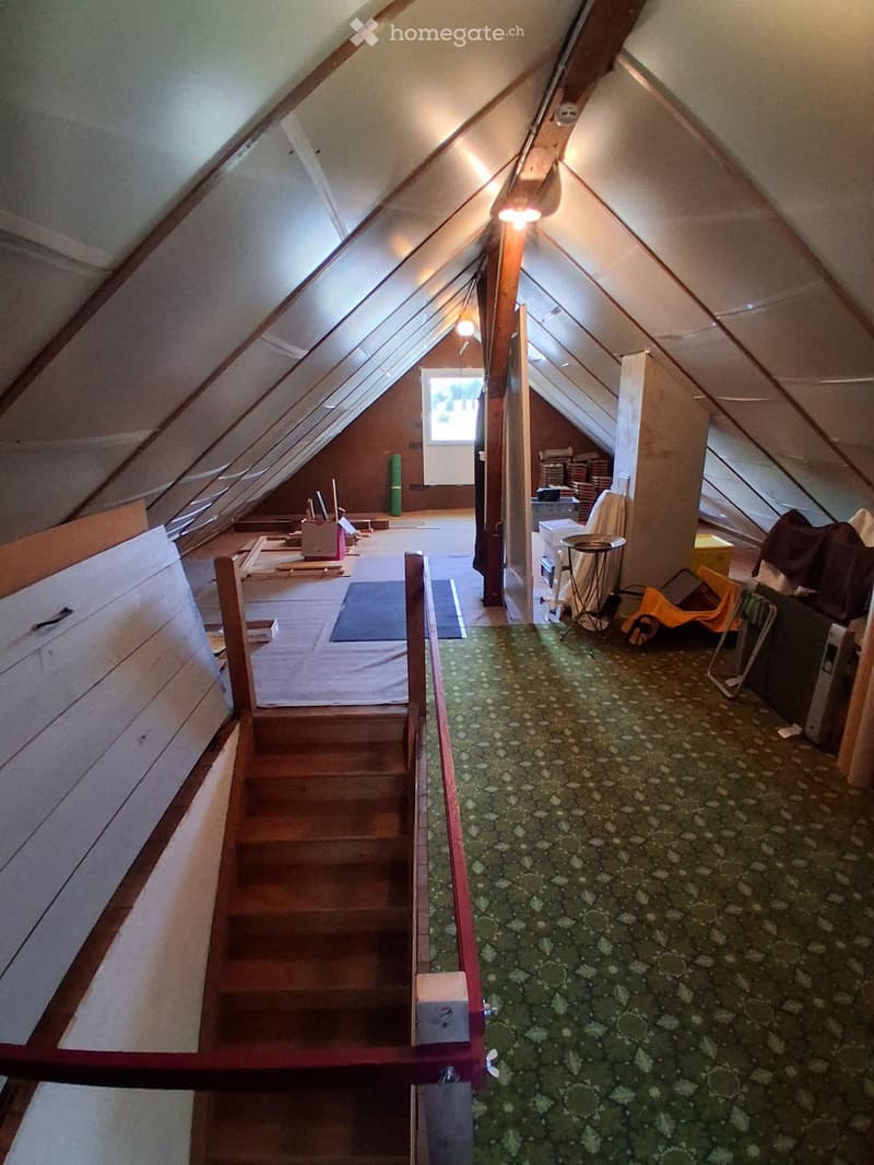 An attic space with exposed wooden beams, a staircase leading down, and a window letting in natural light. The floor is covered with a green carpet, and there are some scattered items including a mattress, a small table, and some storage boxes.