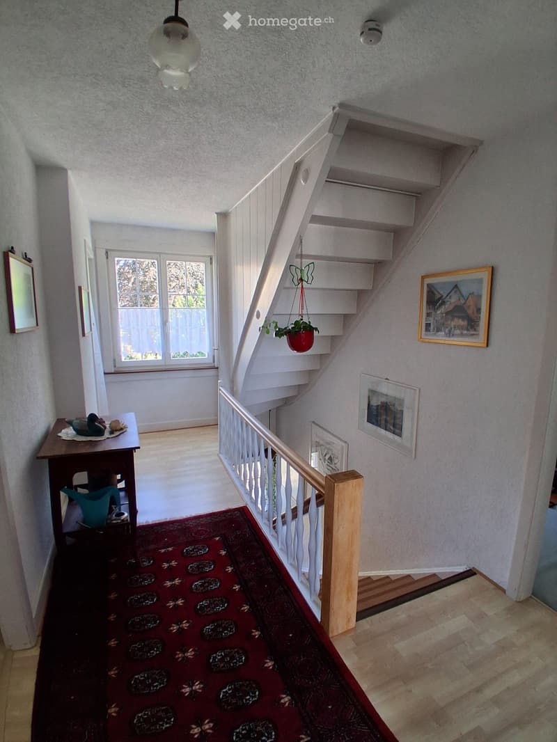 Staircase with white railings, wooden steps, and a hanging plant. A small table with a decorative item sits near the window at the end of the hallway.