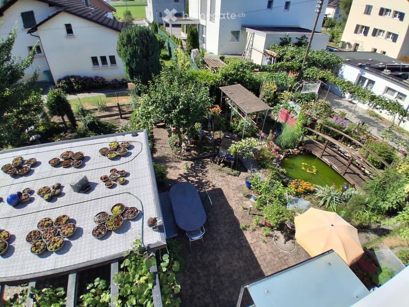 A lush garden with a small pond, wooden bridge, potted plants, and outdoor furniture including a table and chairs.