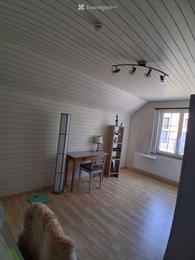 Bright attic room with wooden walls and floor, featuring a desk with a chair, a small shelf, a lamp, and a radiator under the window.