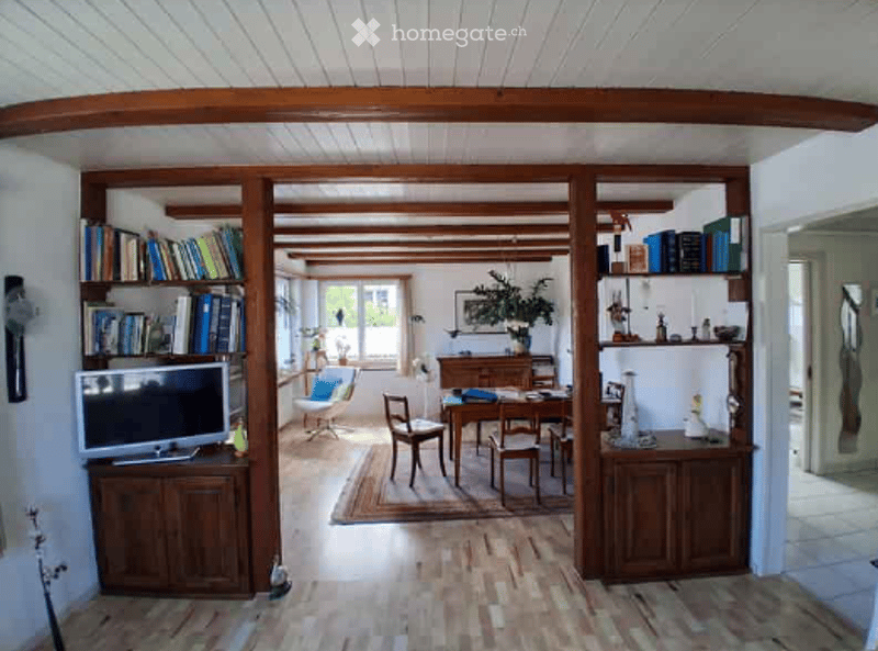 A spacious, open-plan living and dining area with wooden beams on the ceiling, wooden flooring, and large windows. The living area features a sofa, a television on a wooden stand, and a bookshelf filled with books. The dining area includes a wooden dining
