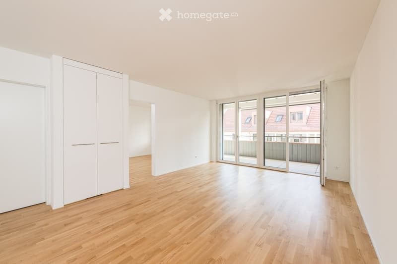 Empty room with light wooden flooring, white walls, large sliding glass doors leading to a balcony, and built-in white closet units.