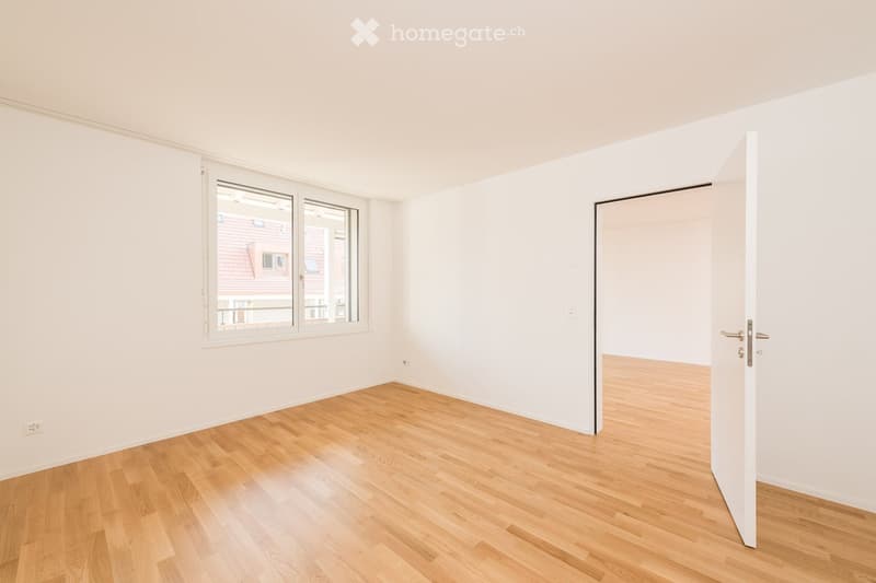 Empty room with light wooden flooring, white walls, and a large window showing a view of neighboring buildings. An open door leads to another space.