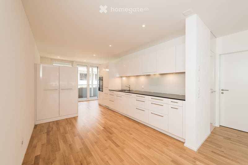 Modern kitchen with white cabinets, wooden flooring, and a sleek sink area. Features include built-in lighting and a sliding glass door leading to a balcony.