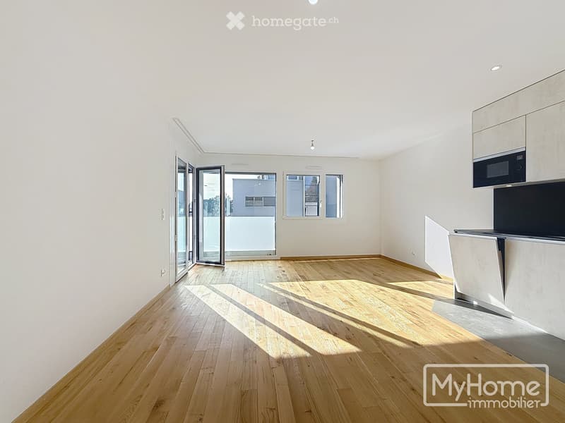Empty room with light wooden flooring, white walls, and large windows allowing natural light. Features a modern kitchen area with cabinets and a microwave, and a sliding glass door leading to a balcony.