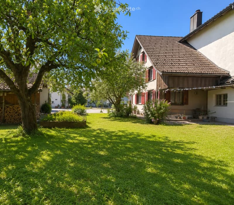 Lush green lawn with a large tree, a wooden house with red shutters, and a stack of firewood on the left side.