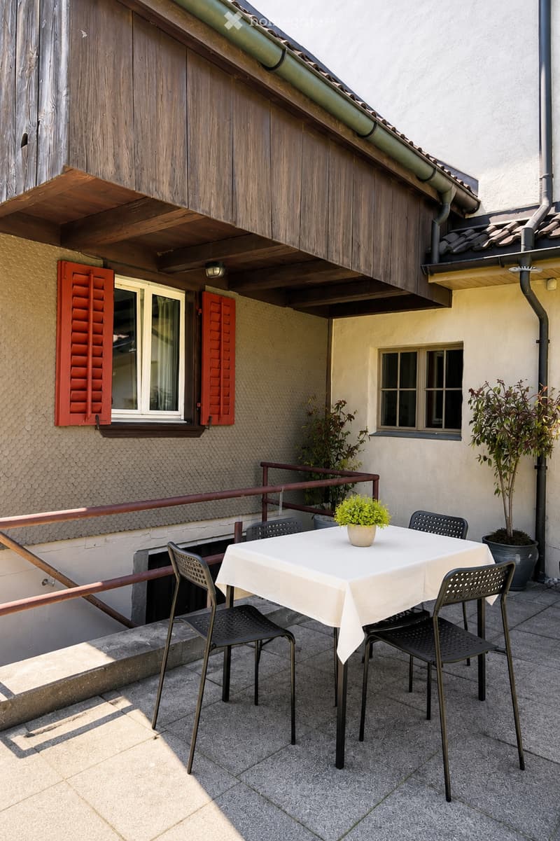Balcony with a white table, four black chairs, and potted plants, adjacent to a building with wooden and white walls, red shutters, and a wooden overhang.