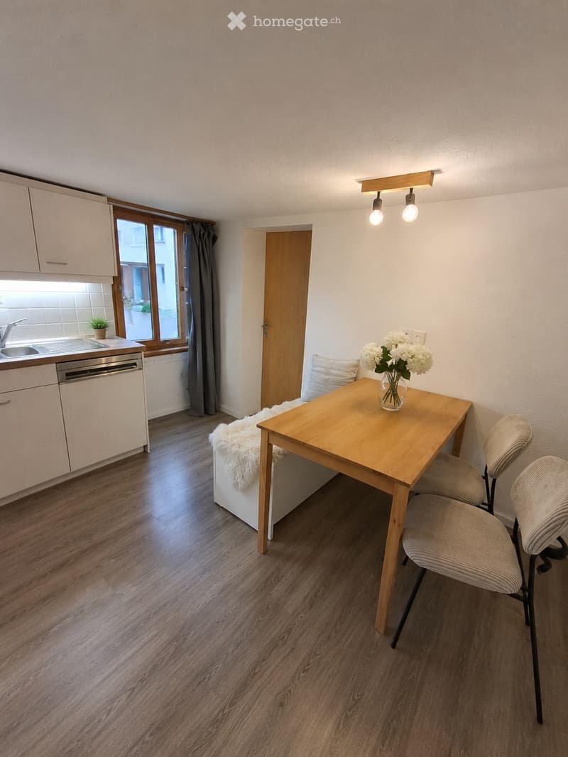 A modern, compact living space with a wooden dining table, chairs, a small sofa, and a kitchen area featuring cabinets, a sink, and a dishwasher. The room has wooden flooring, large windows with curtains, and a decorative flower vase on the table.