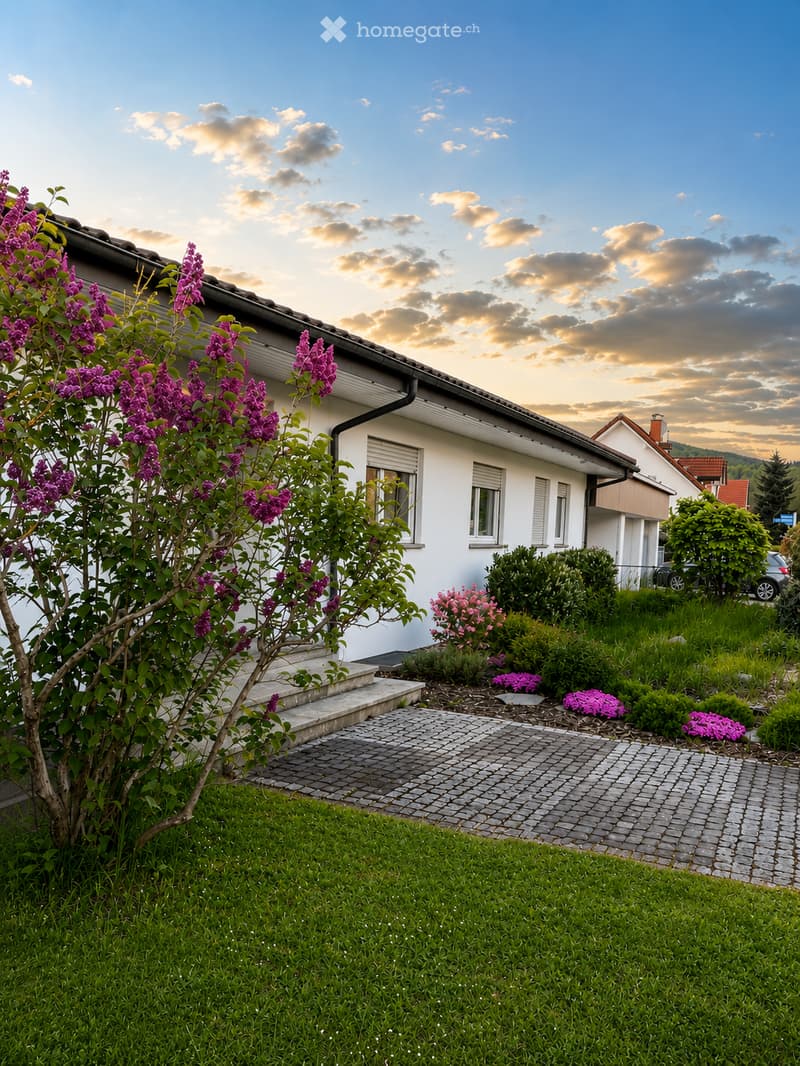 A two-story house with white walls, multiple windows, and a well-maintained garden featuring various flowering plants and shrubs.