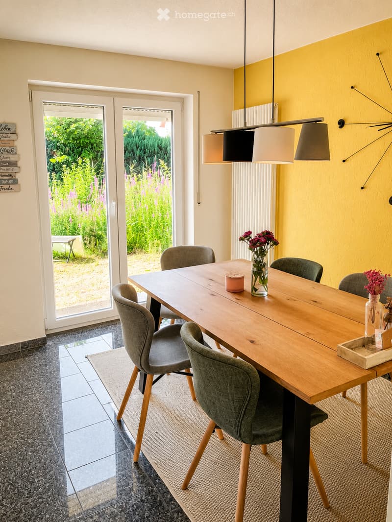 Bright dining room with a large wooden dining table surrounded by modern grey chairs, a vase with flowers, and a view of a lush garden through large glass doors.