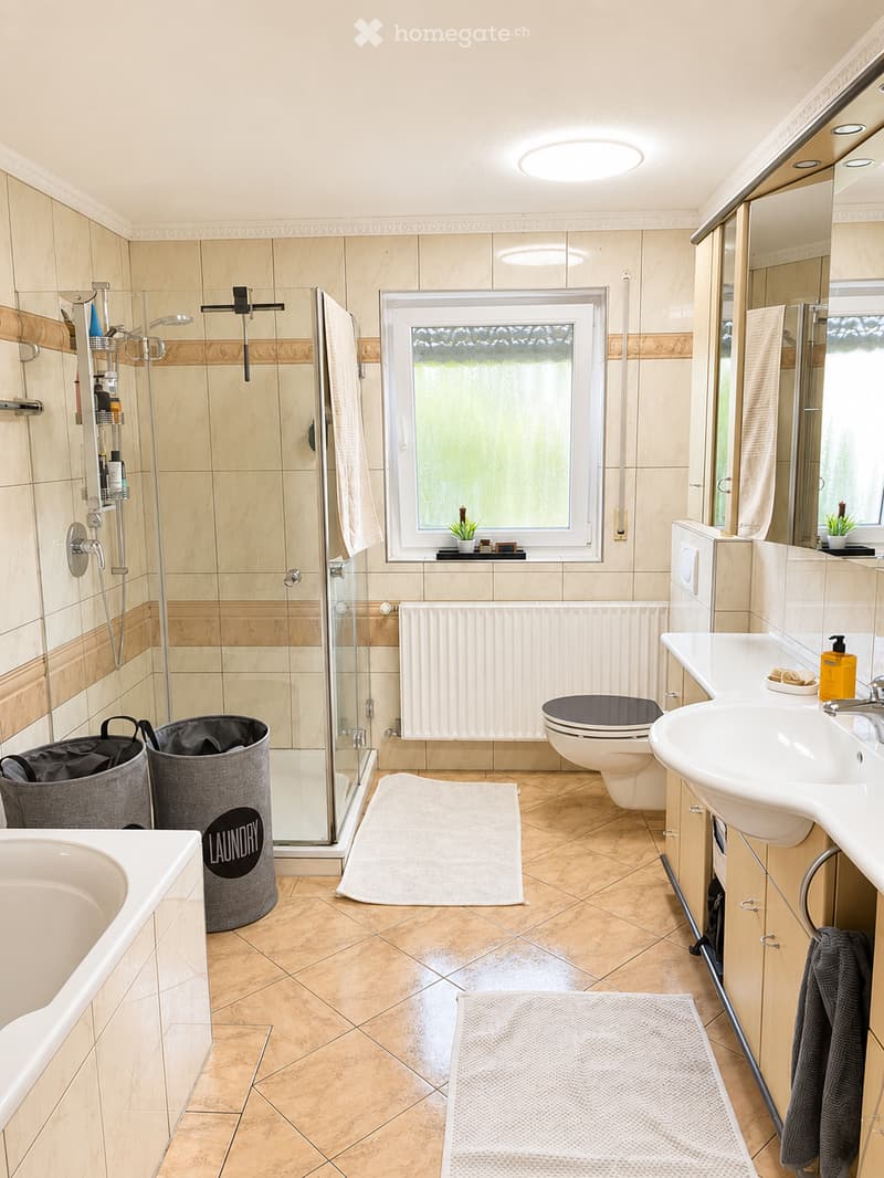 Modern bathroom with beige tiles, a bathtub, toilet, sink, shower, radiator, and two towels on the floor. There is a window with a small plant on the sill and a trash bin near the shower area.