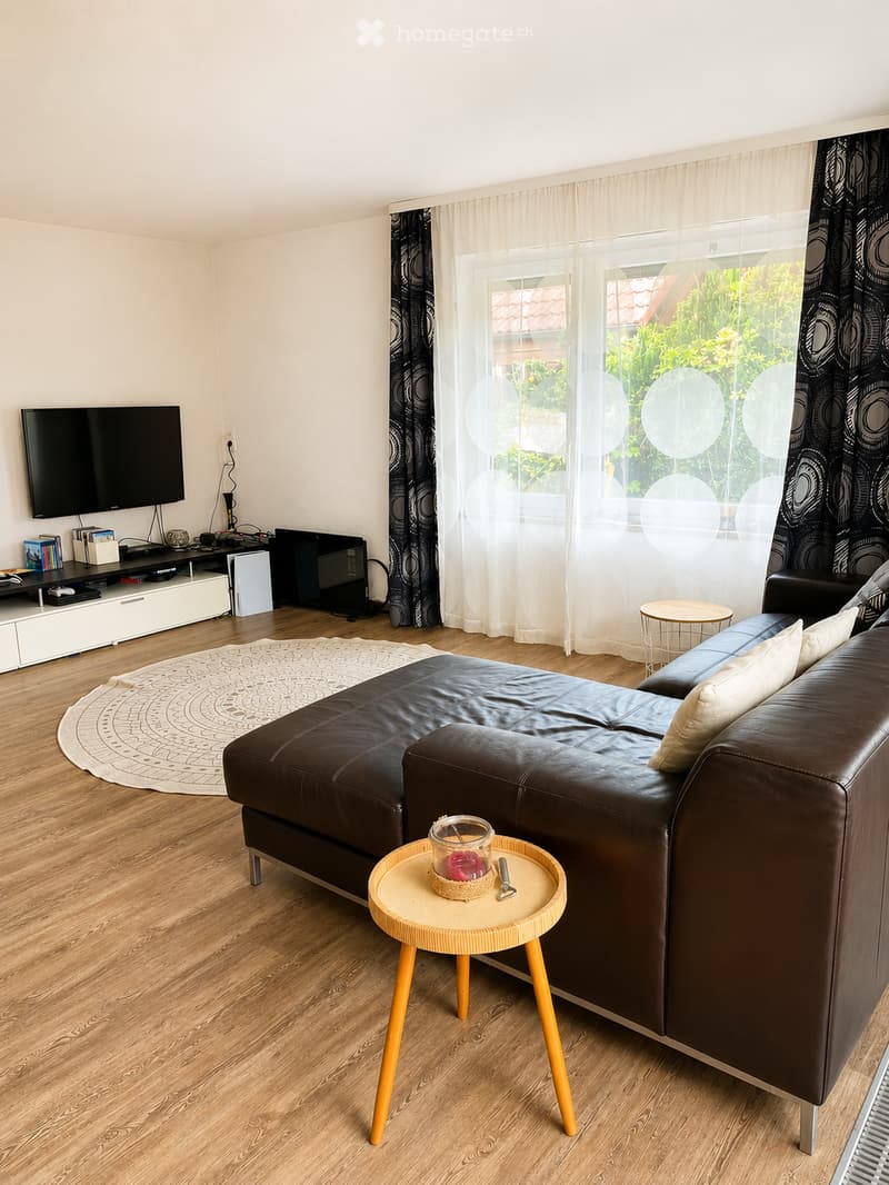 Modern living room with a dark leather sofa, a small round coffee table, a television on a stand, and large windows with curtains.