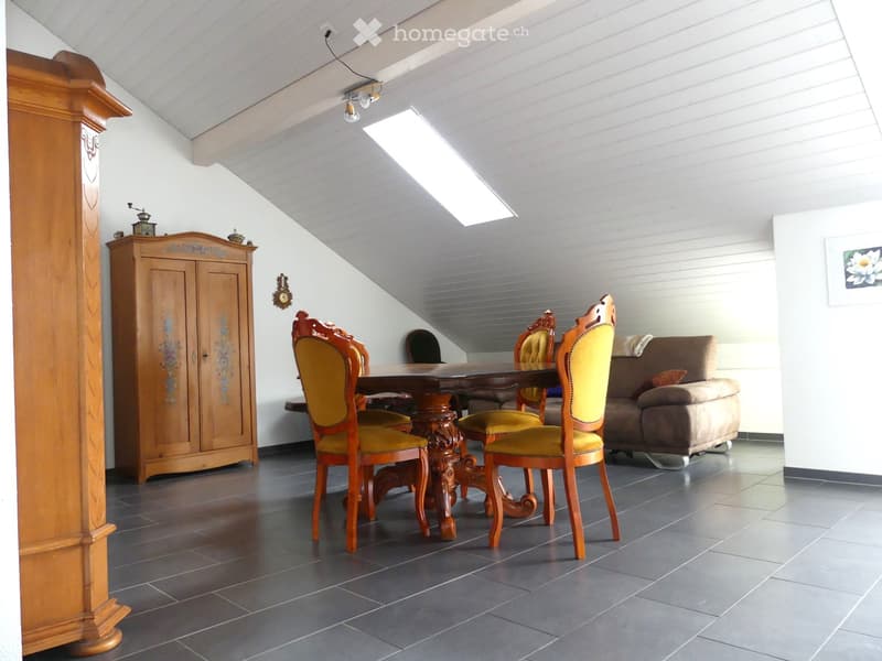 A cozy attic room with a slanted ceiling, featuring a wooden dining table with four ornate chairs, a brown sofa, and a wooden cabinet with floral designs.