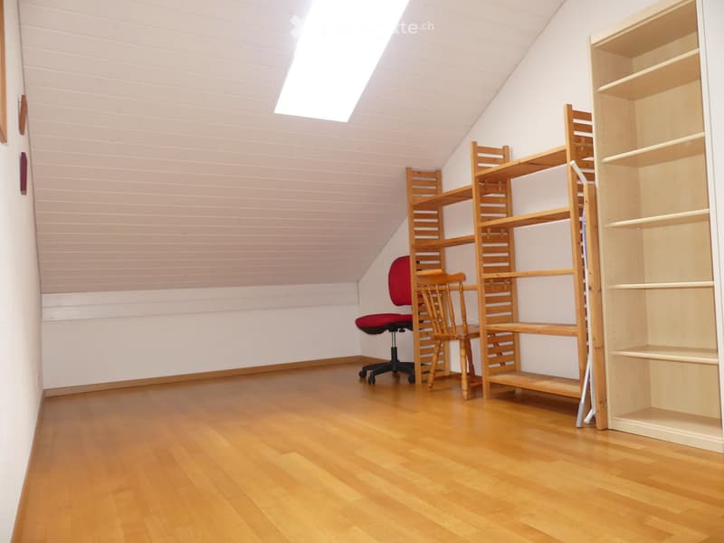 Bright attic room with wooden flooring, slanted ceiling, and skylight. Contains a red office chair, a small wooden table, and built-in wooden shelves.