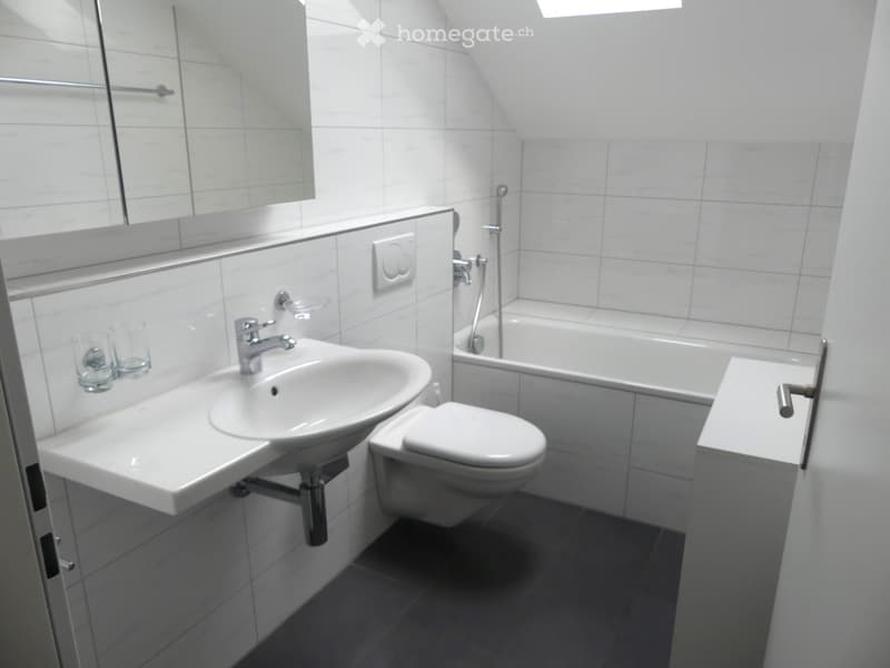 Modern bathroom with white tiles, a sink, toilet, bathtub, and a large mirror. The room features a sleek design with a chrome faucet and a glass shelf holding two cups.