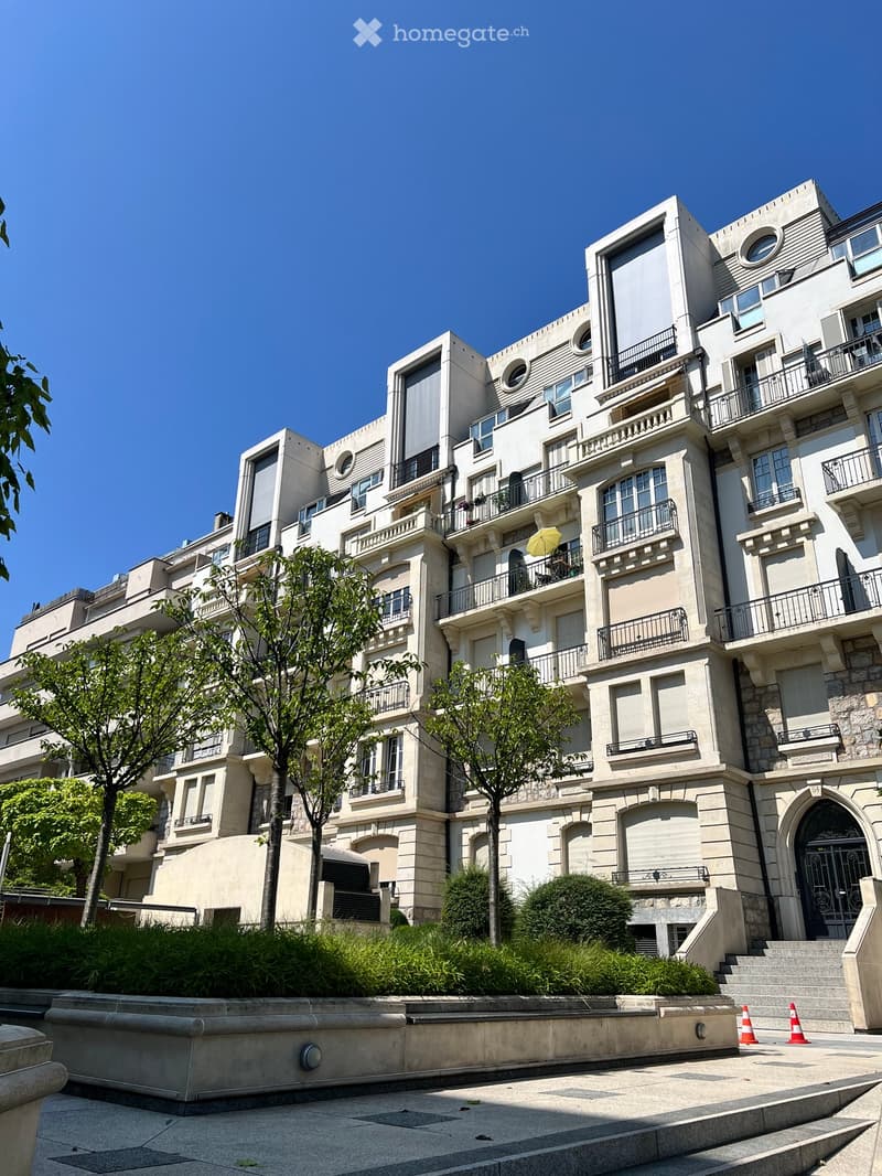 Multi-story residential building with balconies, large windows, and ornate stonework. Trees and shrubs are planted in front of the building.
