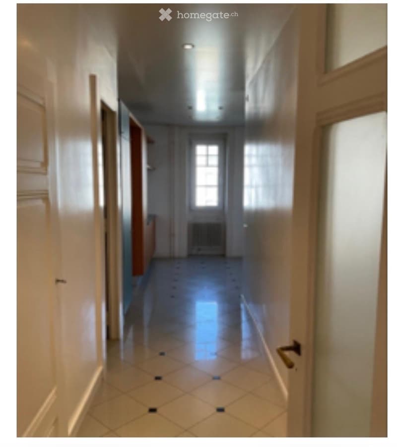 A long, narrow hallway with tiled flooring, white walls, and several doors on both sides. Natural light streams in from a window at the far end, and the ceiling features recessed lighting.
