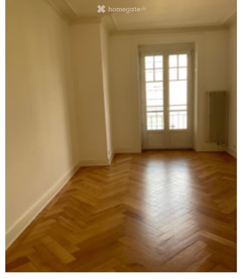 Empty room with wooden herringbone flooring, light-colored walls, and a large window with multiple panes allowing natural light to enter.