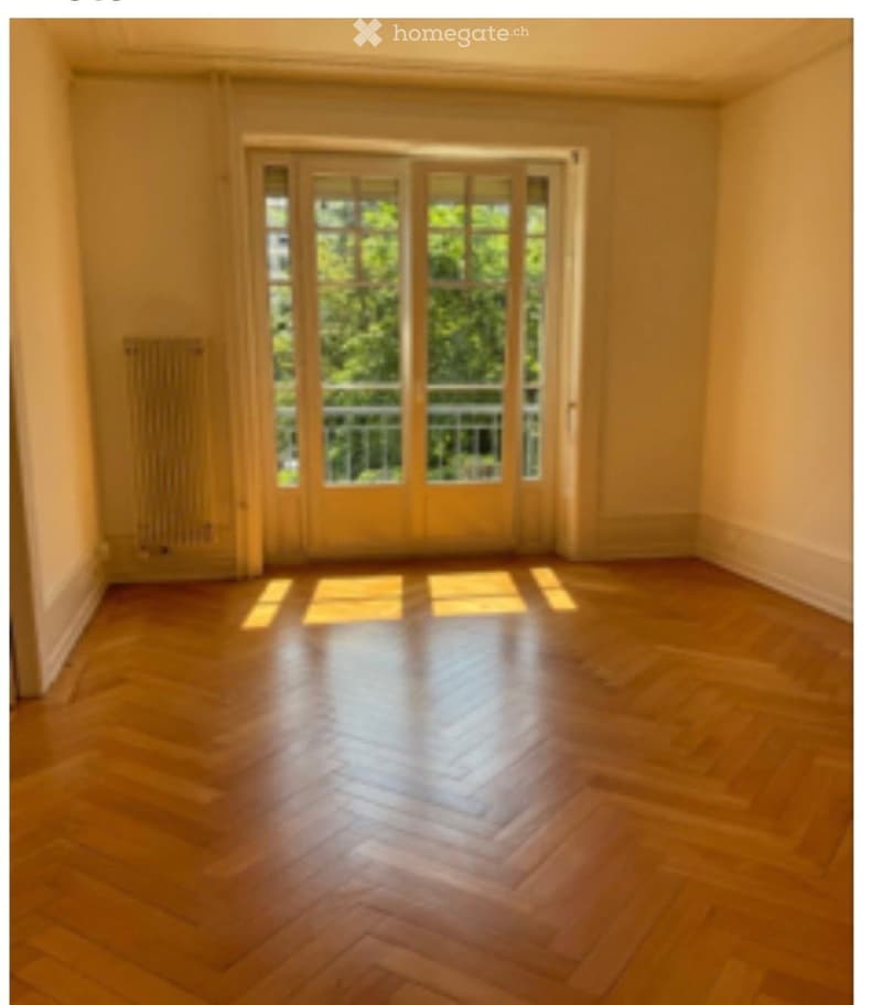 Empty room with wooden flooring and large glass doors leading to a balcony. The walls are painted in a light color, and the room is well-lit with natural light.